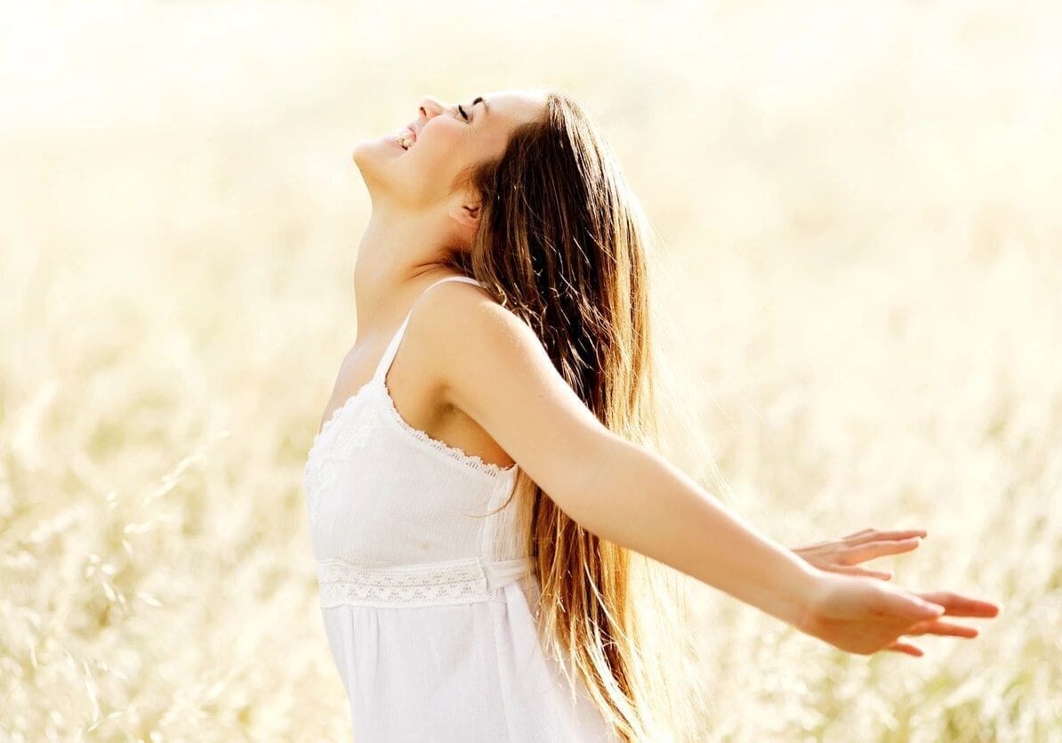 A woman in a white dress stands in a bright, open field, tilting her head back and extending her arms outward.